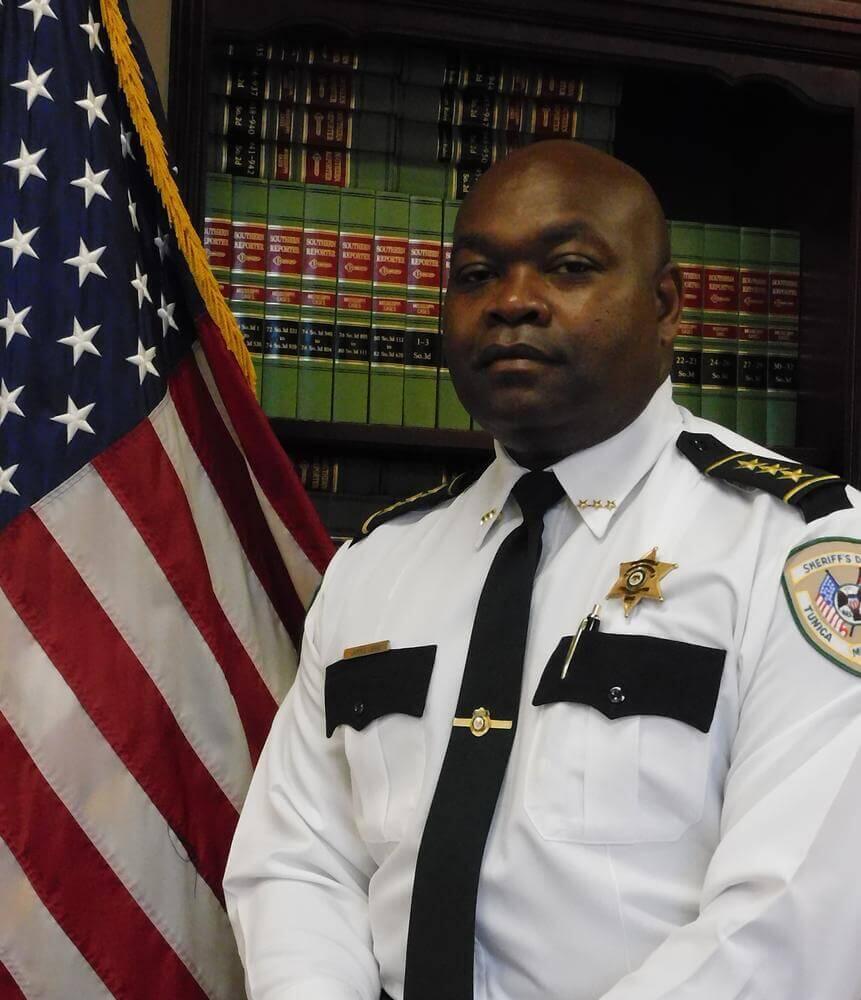 Assistant Chief Deputy Jammie Lewis standing next to the Flag of the United States of America.